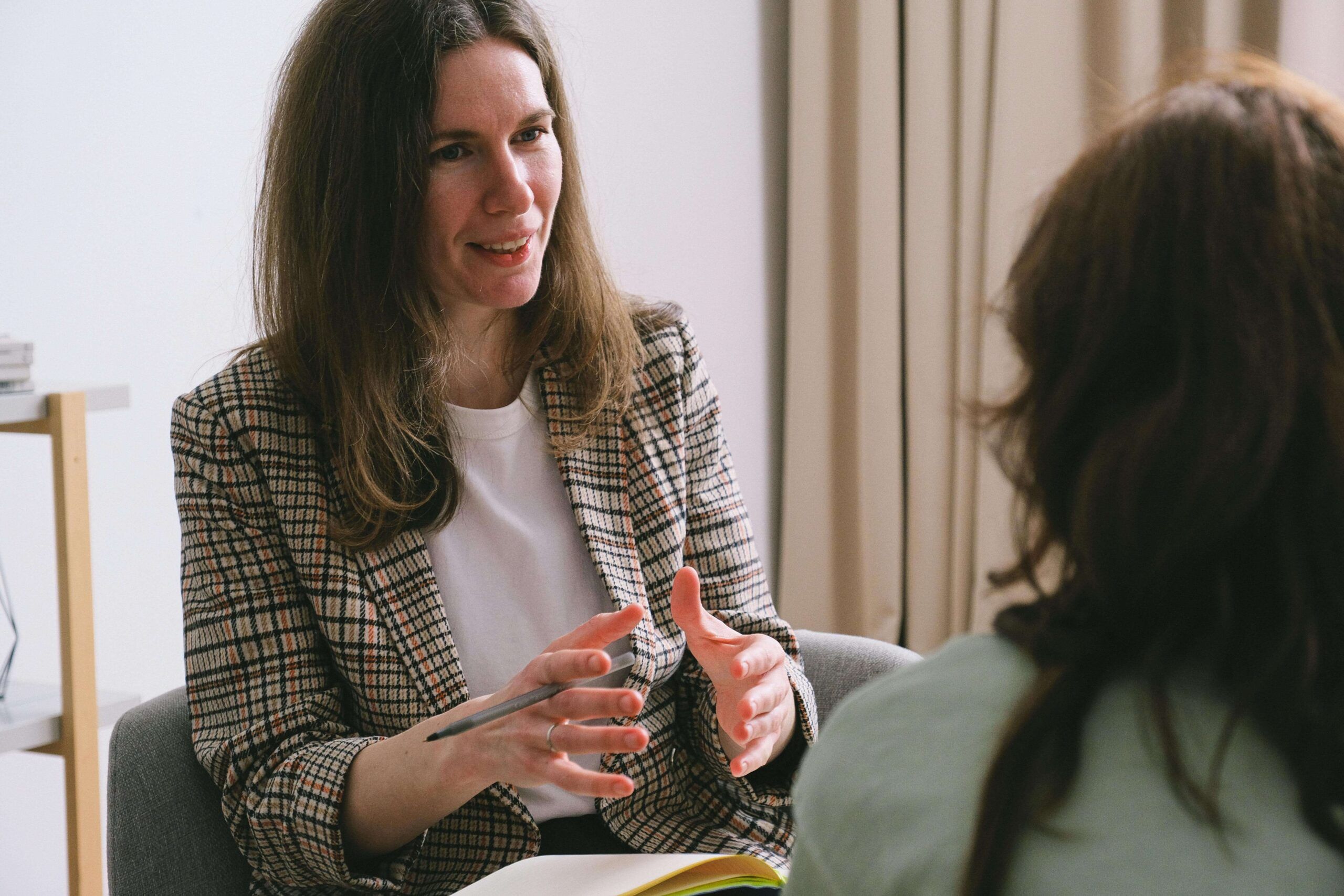 Two women engaged in a thoughtful conversation, one gesturing with a pen while smiling warmly. Perfect for a blog on finding a nutritional therapist in Raleigh, NC, and the benefits of nutritional counseling in Raleigh, NC, for transformative health changes.