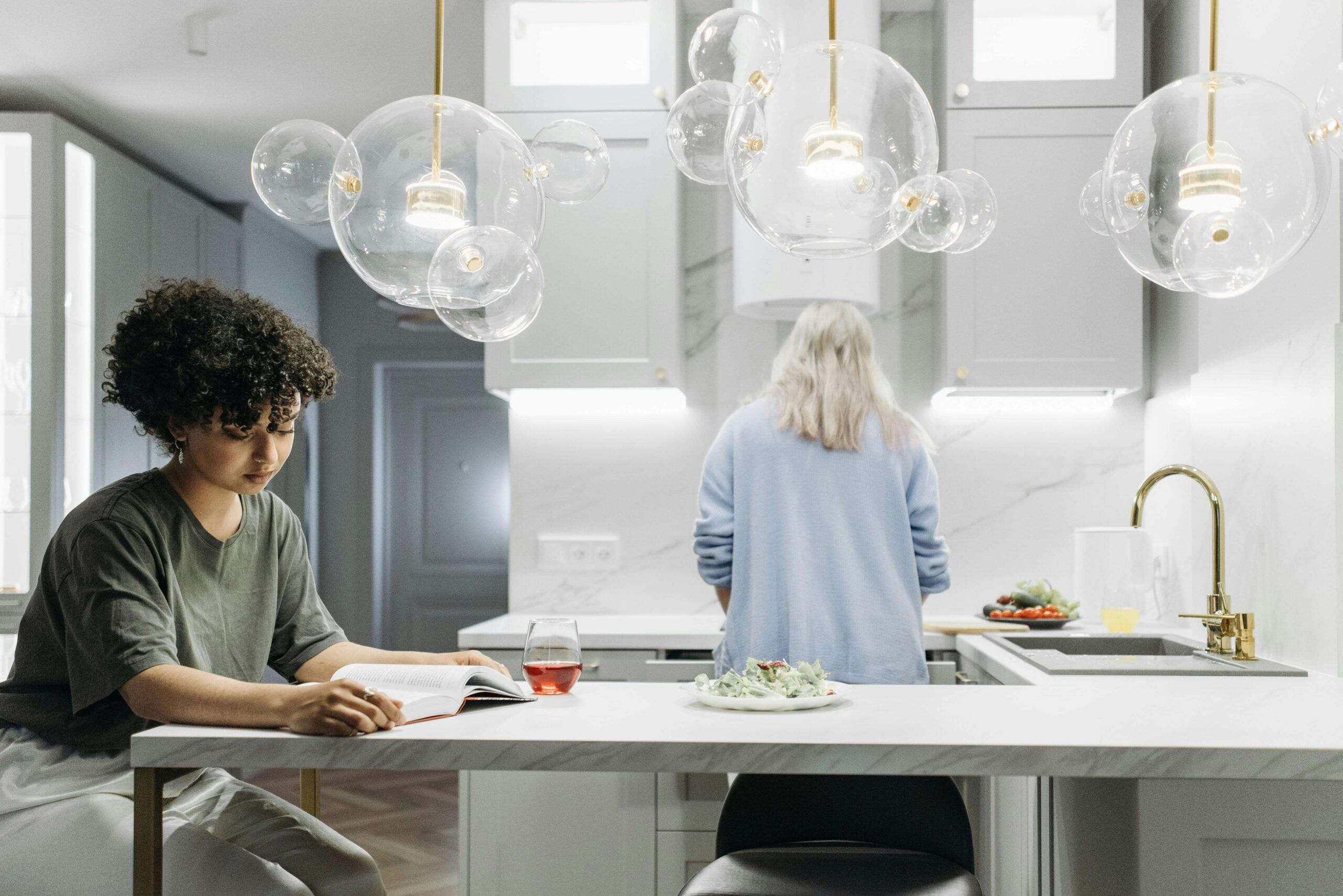 A woman reads at a kitchen counter while another prepares food in the background, symbolizing reflection and support after GLP-1 use with a nutritional therapist in Raleigh, NC offering compassionate nutritional counseling in Raleigh, NC for post-medication care.