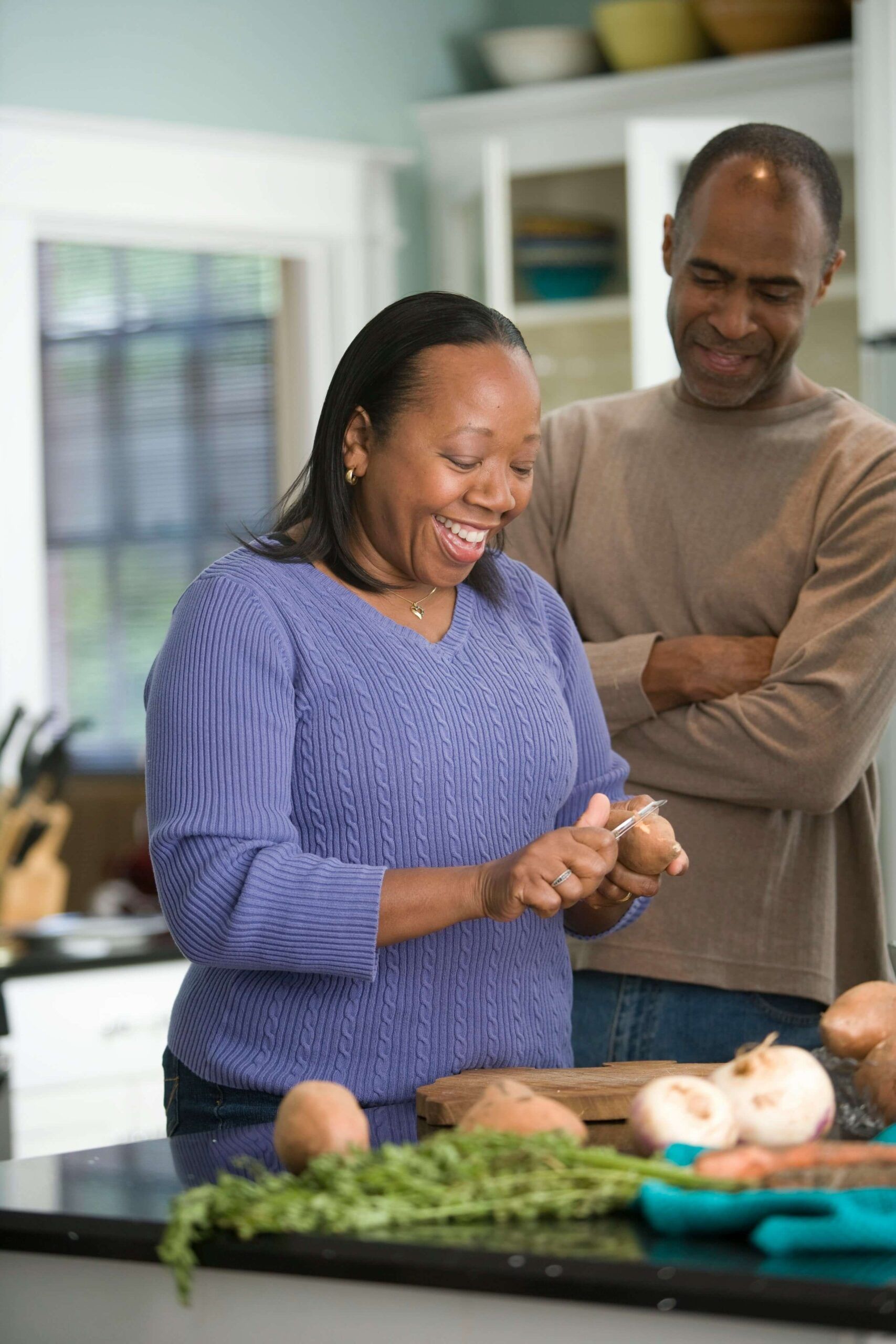 A joyful middle-aged woman peels root vegetables at a kitchen counter while a man stands beside her smiling, representing the support and guidance available through nutrition counseling in Raleigh, NC. Learn how a registered dietitian in Raleigh, NC can help you feel grounded and empowered during menopause.