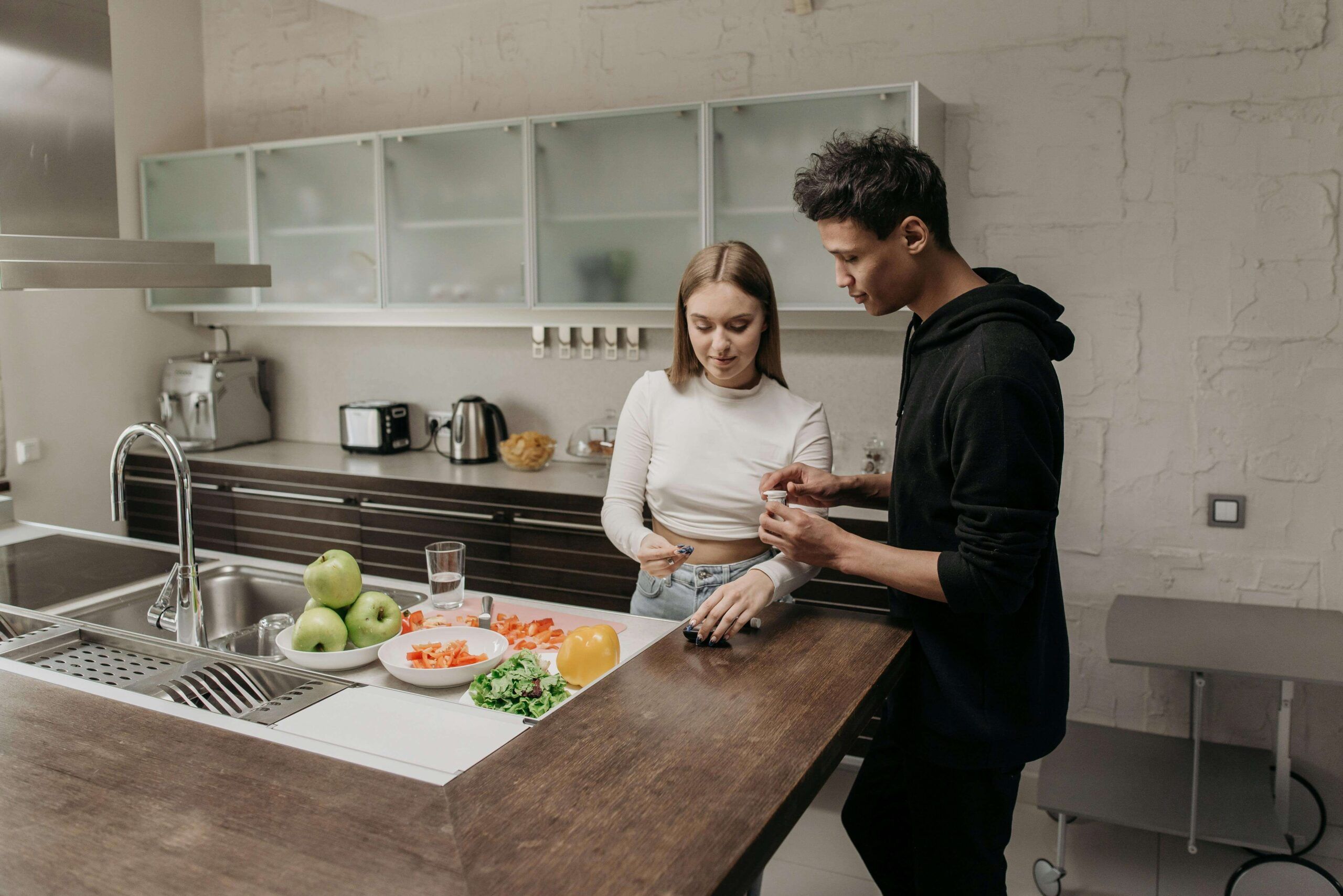 A young couple preparing food together at home, surrounded by fresh vegetables, while checking a glucose monitor—representing support through nutritional counseling in Raleigh, NC with guidance from a nutritional therapist in Raleigh, NC.