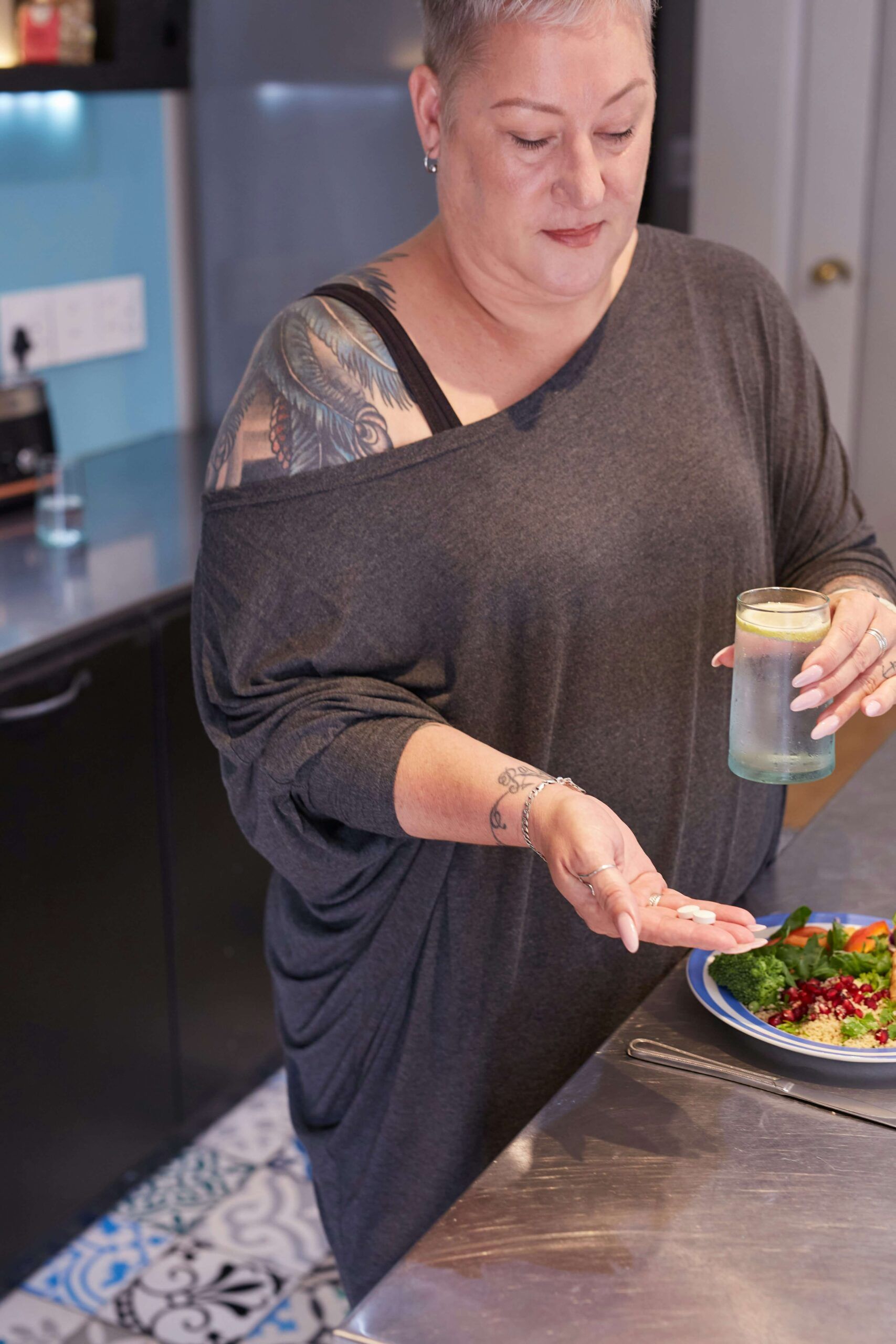 A woman with short hair and tattoos holds a glass of water and supplements while standing near a colorful salad, representing support from a registered dietitian in Raleigh, NC through nutritional counseling in Raleigh, NC for managing chronic conditions with personalized care.