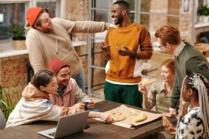 A diverse group of friends sharing pizza and laughing around a table, highlighting the social and emotional aspects of eating. Support from a registered dietitian in Raleigh, NC or through binge eating disorder therapy Raleigh, NC can help you feel more at ease in food-related settings and rebuild trust in your relationship with food.