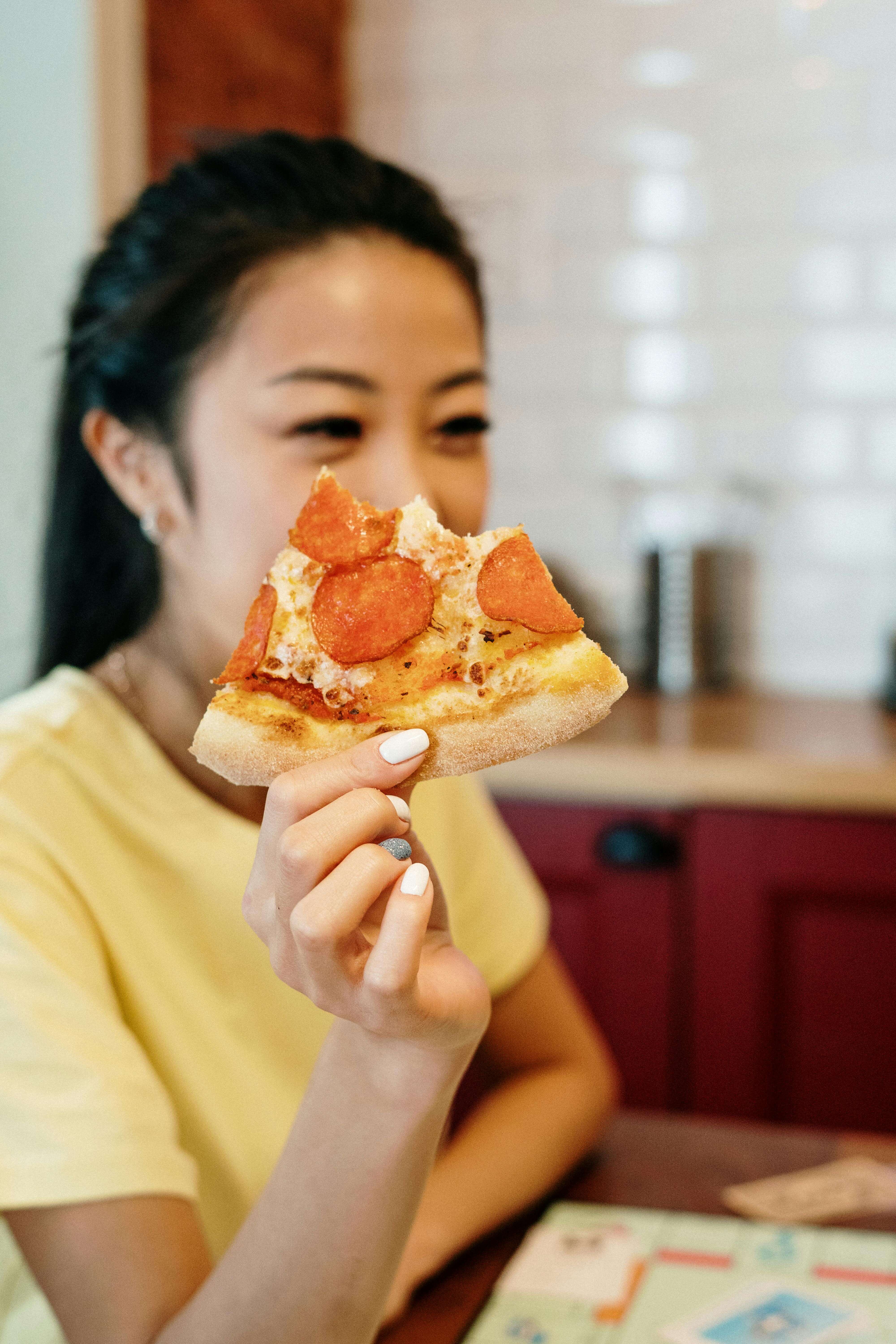 A young woman holds a slice of pepperoni pizza while smiling, illustrating the freedom and satisfaction supported through nutrition therapy Raleigh, NC and nutritious counseling for eating disorders Raleigh, NC.