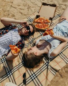 Two people relax on a beach blanket enjoying pepperoni pizza in the sun, reflecting joyful eating and food freedom supported by nutritional counseling in Raleigh, NC and nutritious counseling for eating disorders Raleigh, NC.