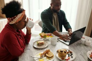 A couple enjoying a mindful breakfast together at a kitchen table, with one partner sipping coffee and the other working on a laptop, representing how working with a nutritional therapist in Raleigh, NC and receiving support through nutrition counseling with a registered dietitian in Raleigh, NC can support intuitive eating during menopause.