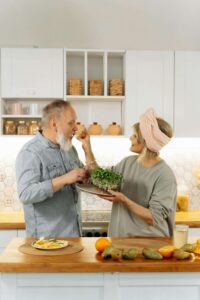 Older couple enjoying time together in a bright kitchen, with one feeding the other a sprout while surrounded by fresh produce—highlighting the joy of nourishing habits during menopause with support from nutritious counseling in Raleigh, NC and eating disorder therapy in Asheville, NC.