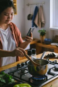 A woman stirs a pot on the stove while checking her phone, capturing the mental load that comes with trying to figure out nourishment alone — nutrition counseling in Raleigh, NC with a registered dietitian in Raleigh, NC can help carry that weight.