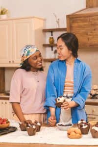 Two women laugh together in a warm kitchen surrounded by cupcakes and fresh fruit, capturing the joy and ease around nourishment that medical nutrition therapy in Raleigh, NC and a nutritional therapist in Raleigh, NC can help make possible.
