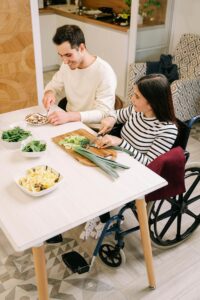 A woman using a wheelchair and a man prepare a meal together at a kitchen table with fresh vegetables and pasta, reflecting the everyday nourishment experiences supported through nutrition counseling for eating disorders in Raleigh, NC and with a dietitian in Hendersonville, NC.