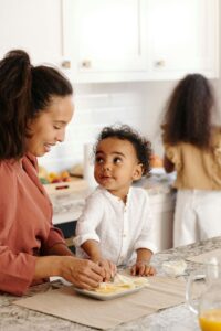  A mother smiles at her toddler while preparing food at a kitchen counter, with another child playing nearby, reflecting the everyday nourishment moments supported through nutrition counseling for eating disorders in Raleigh, NC and with a dietitian in Hendersonville, NC.