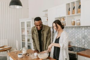 A man and woman laugh together while whisking batter in a bright kitchen, capturing the kind of easy, joyful relationship with nourishing yourself that a nutritional therapist in Raleigh, NC and disordered eating therapy in Raleigh, NC can help make possible.