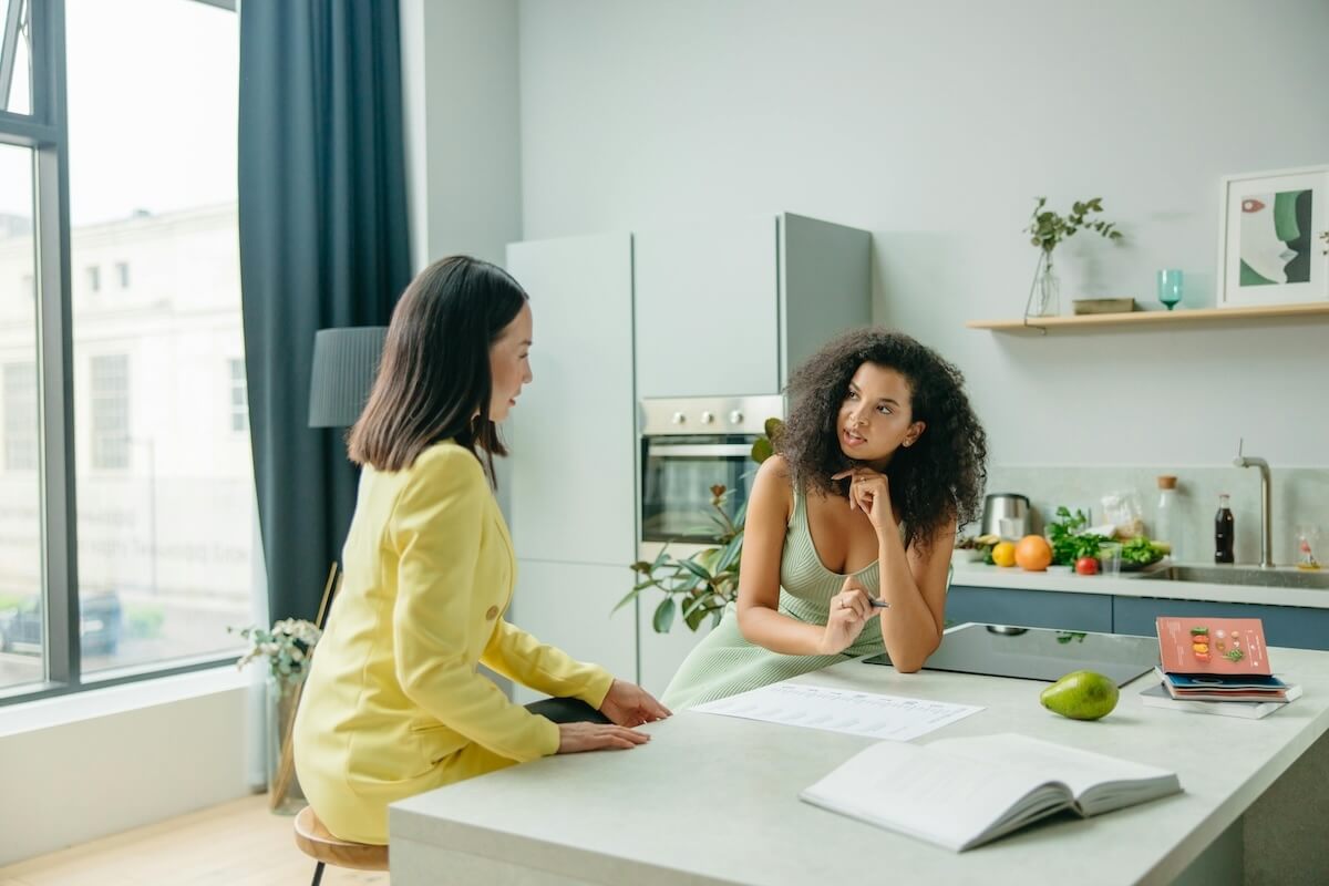 A nutritional therapist in Raleigh, NC sits across from a client at a kitchen counter, listening warmly during a nutrition therapy session in Raleigh, NC, with an open notebook and fresh fruit nearby.